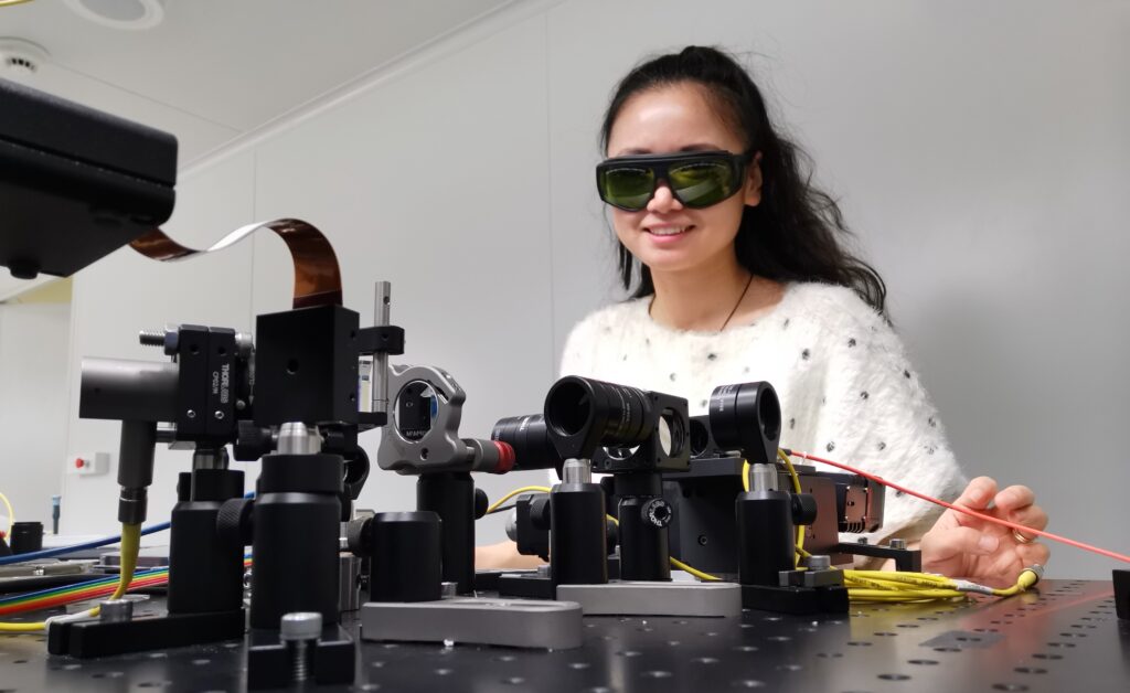 Fiona Wei, wearing dark green laser safety goggles and a white speckled jumper, smiles at the camera from behind a low-angle view of an optical breadboard. In the foreground sit Thorlabs cage-mounted lens tubes, kinematic mirror mounts and fibre collimators, with yellow patch fibres, a red-jacketed fibre, and a ribbon of multicoloured wires running off the table. Her right hand rests near a fibre-coupled component on the right of the bench.